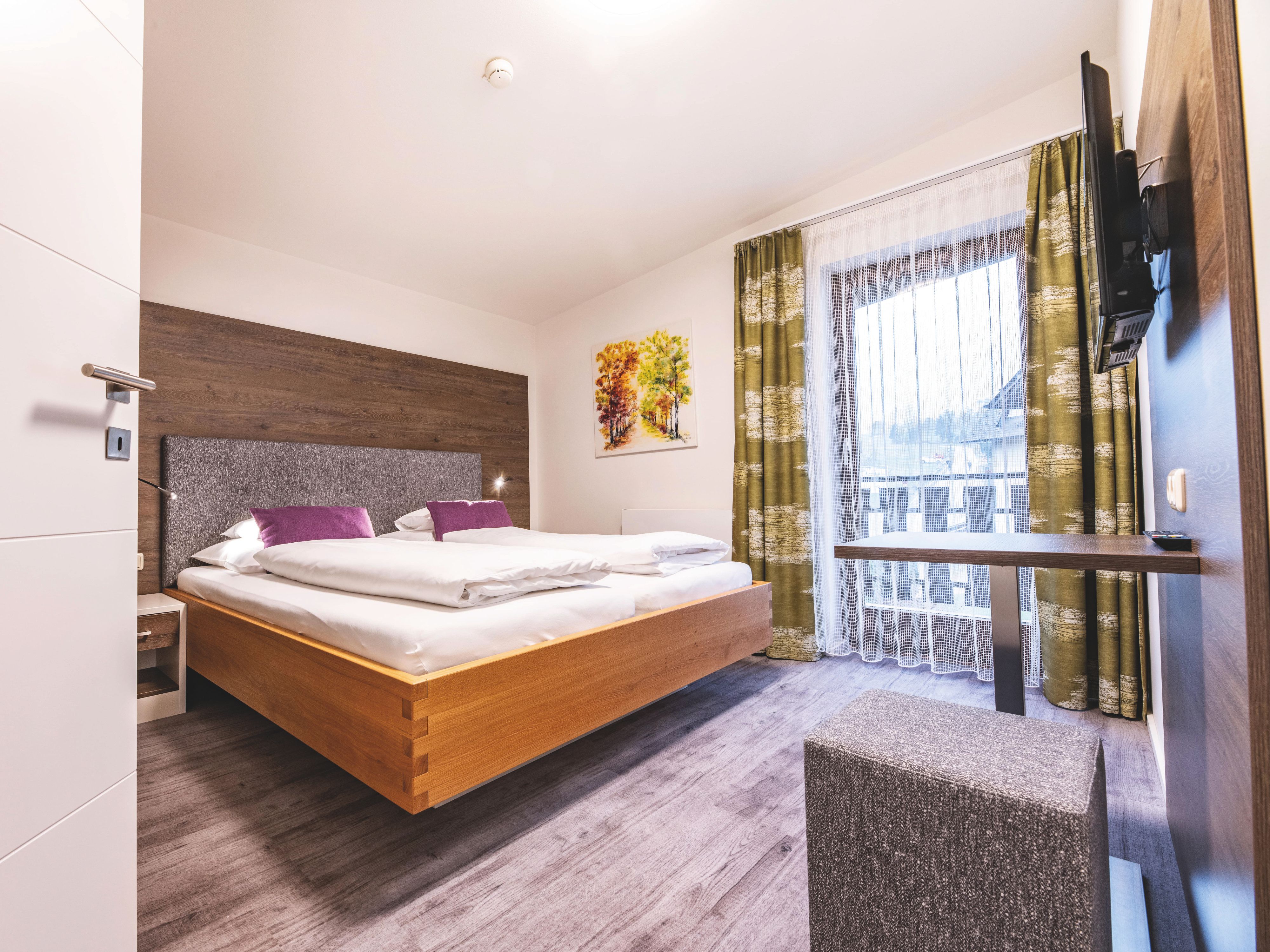 A modern hotel room with a double bed, pillows, wooden furniture, and a balcony.