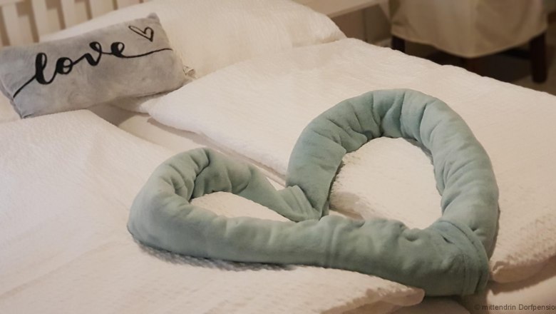 A bed with white pillows and blankets, a gray pillow with the word 'love' on it and a heart-shaped, green fabric pillow.