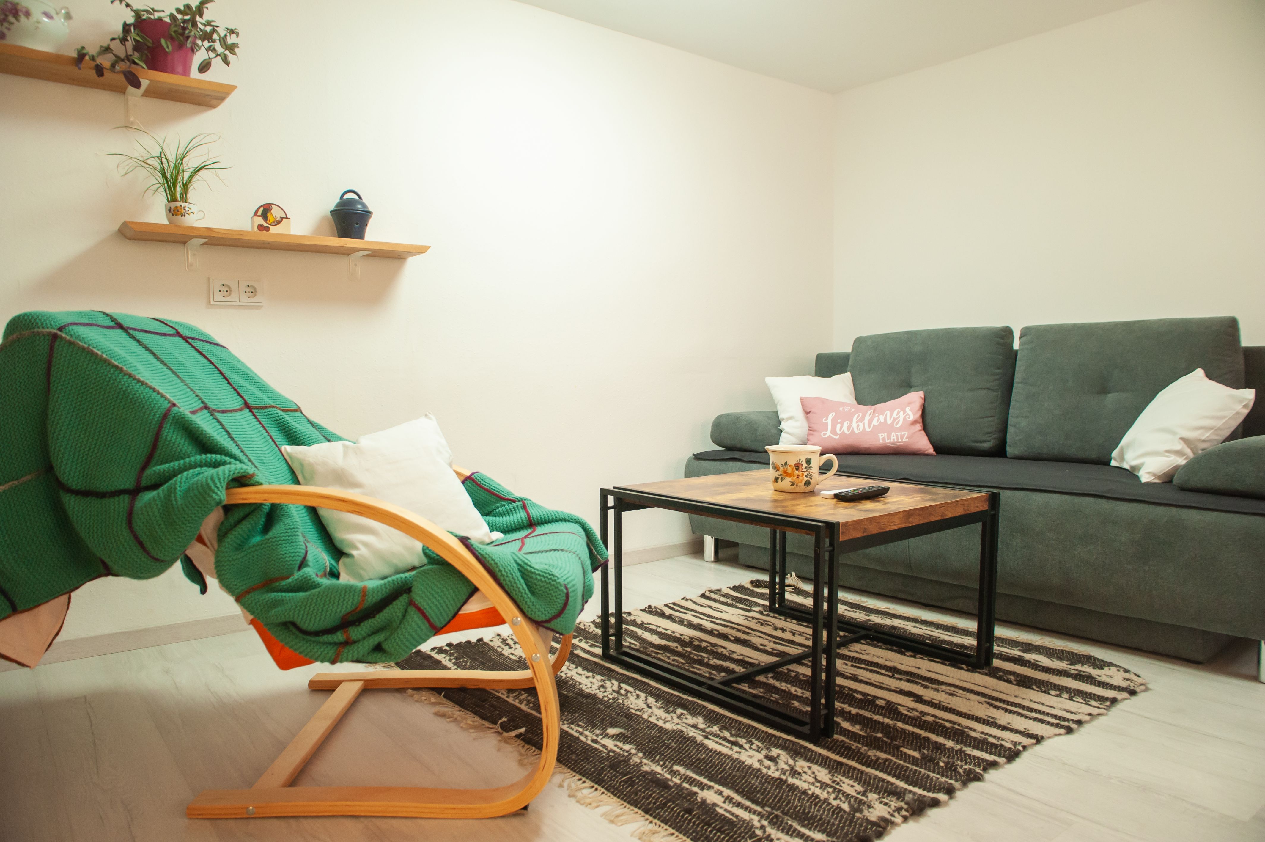 Cozy living room with grey sofa, wooden table, green rocking chair and shelves with plants.