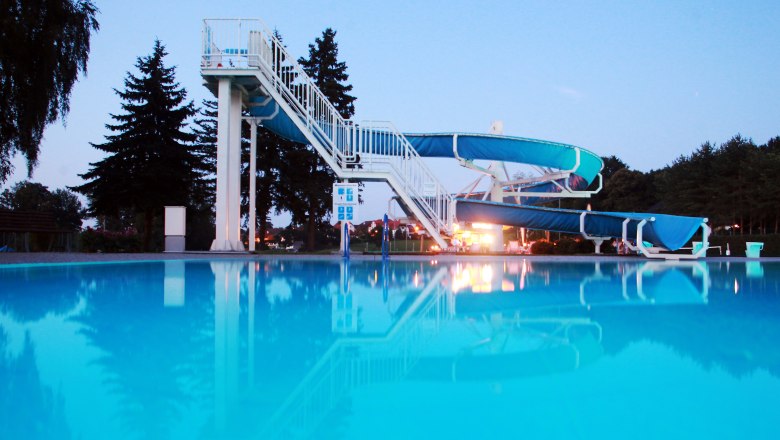 An outdoor pool with a large blue water slide and a clear pool in the foreground, surrounded by trees at dusk.