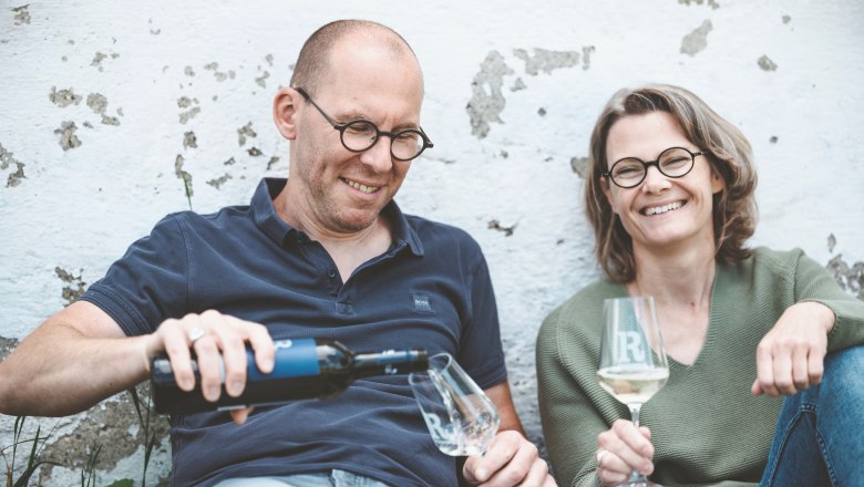 Two people sit relaxed against a wall and drink wine.