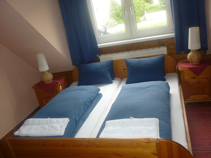 A cozy double room with two single beds, blue bed linen and two bedside tables with lamps, under a window with blue curtains.