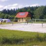 People play volleyball on a sand court in front of a yellow building with a red roof, surrounded by trees.