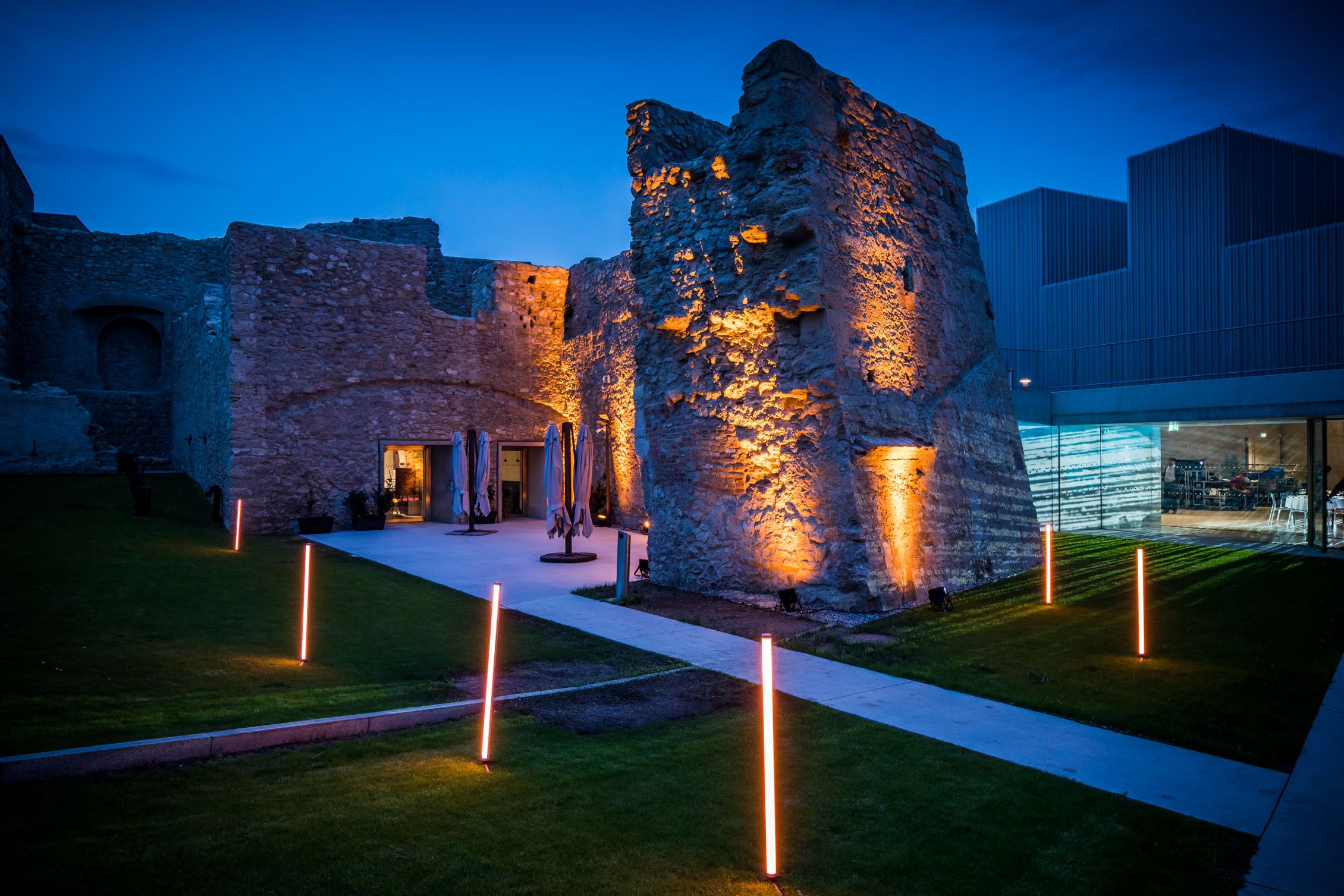 Illuminated ruins with modern buildings in the background at dusk.