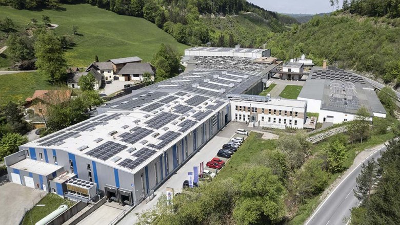 Aerial view of a large factory with solar panels on the roofs, surrounded by a green landscape.