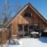 A wooden holiday home in the snow with garden furniture and bare trees.