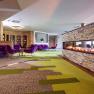 Modern hotel lobby with green-brown carpeting, fireplace, purple sofas and chairs and wooden tables.