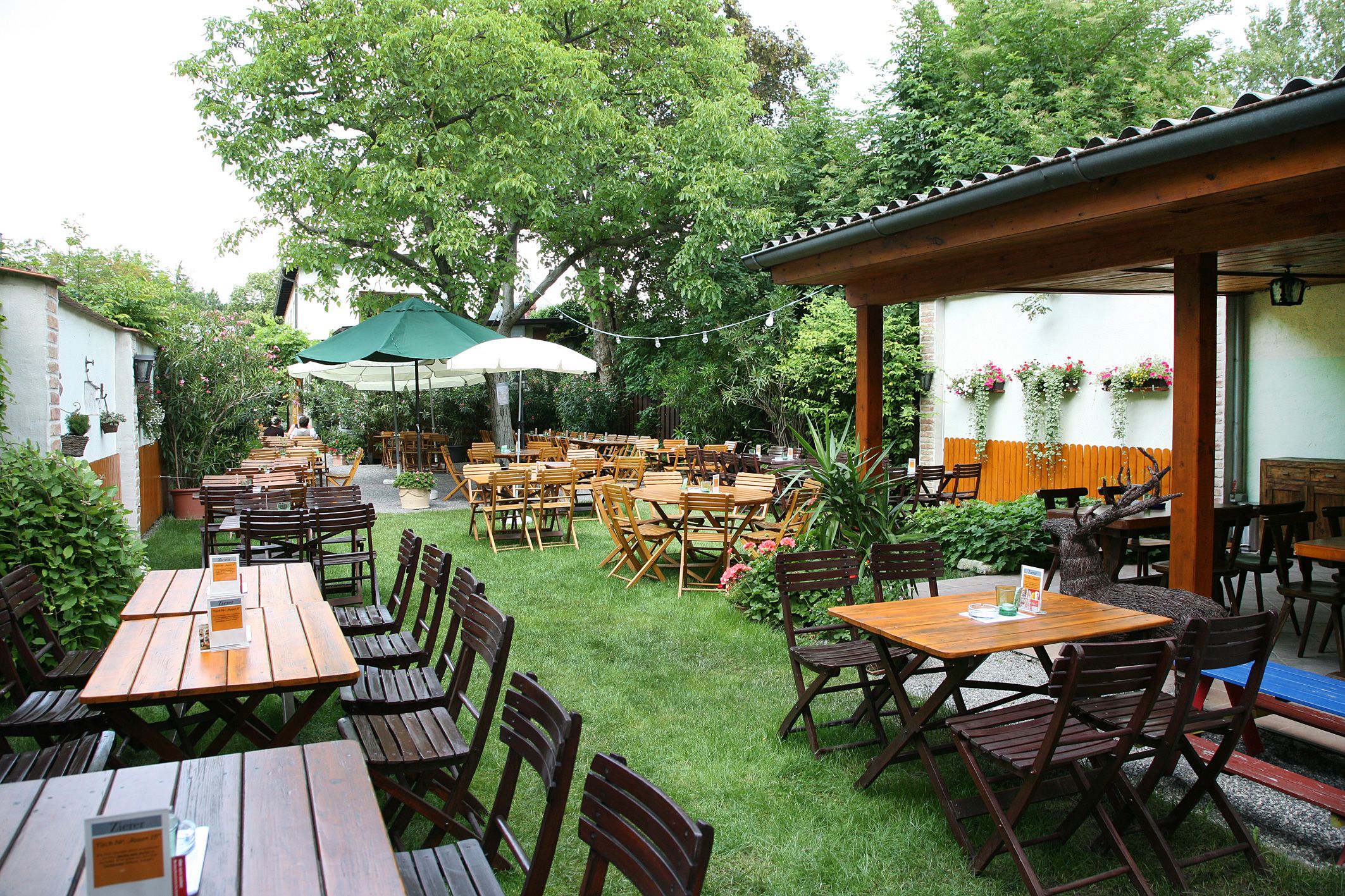 A cozy beer garden with wooden tables and chairs, surrounded by green trees and plants.