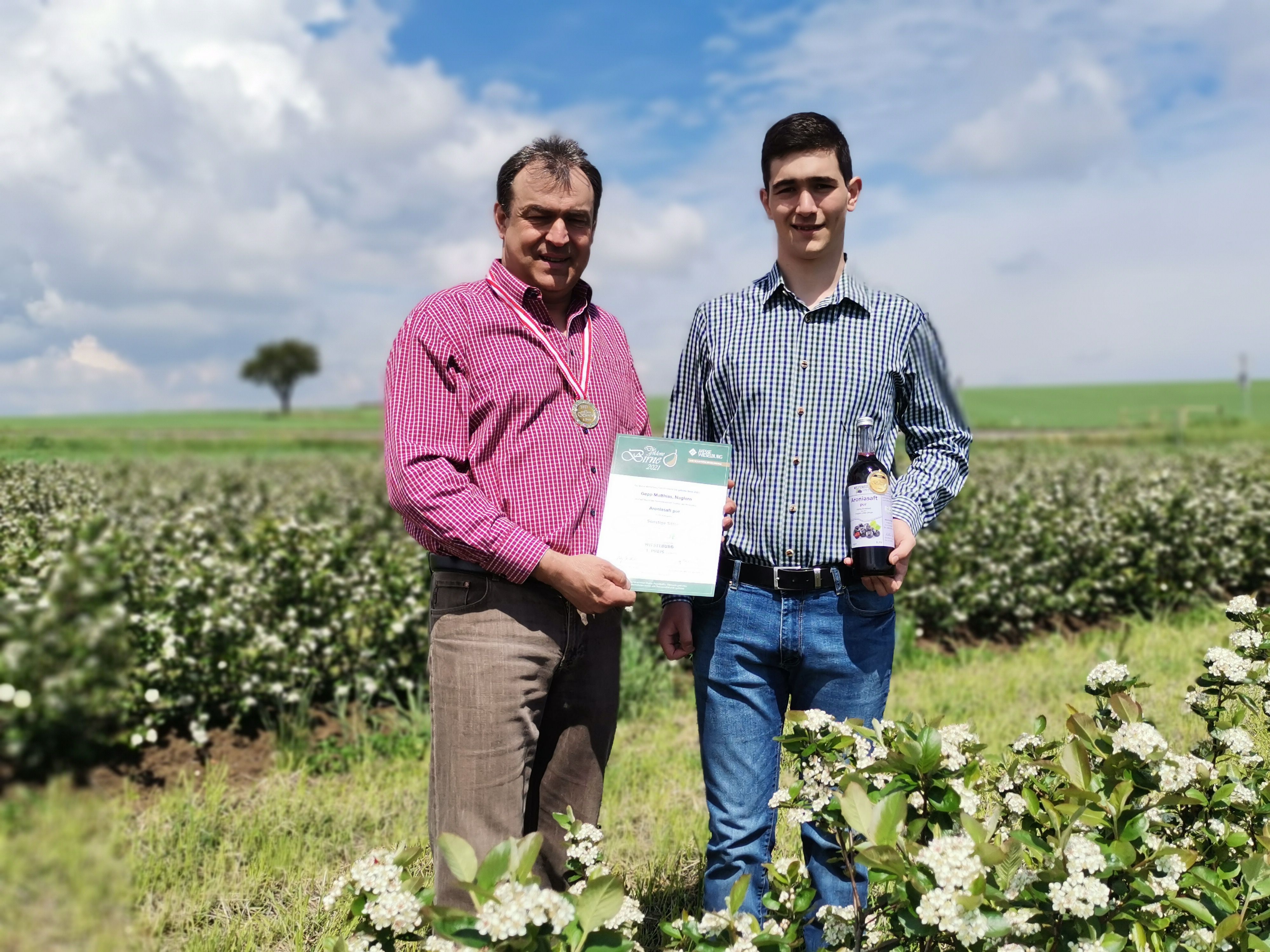 Two men stand in a flowering field, one holding a certificate and a medal, the other a bottle.