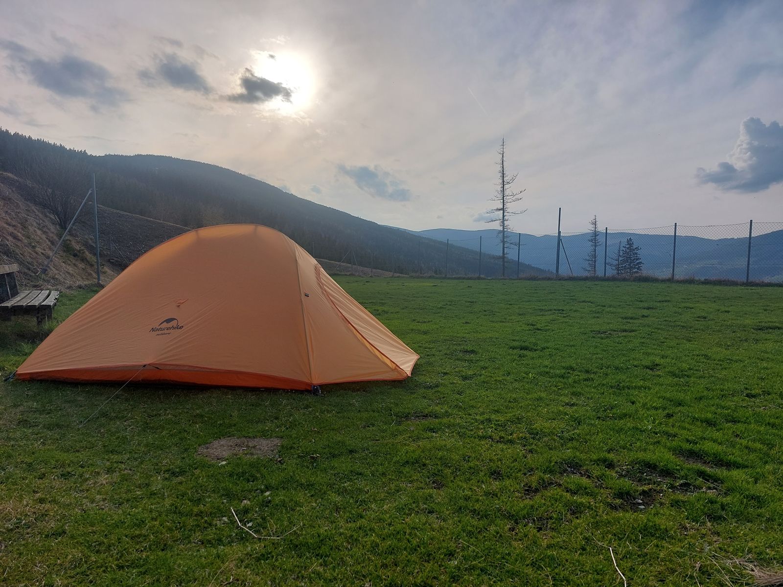 An orange tent stands on a green meadow, surrounded by mountains and a fence, with a cloudy sky and sunset.