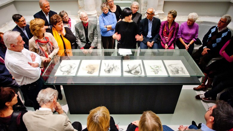 A group of people stand around a glass table with drawings in an exhibition room.