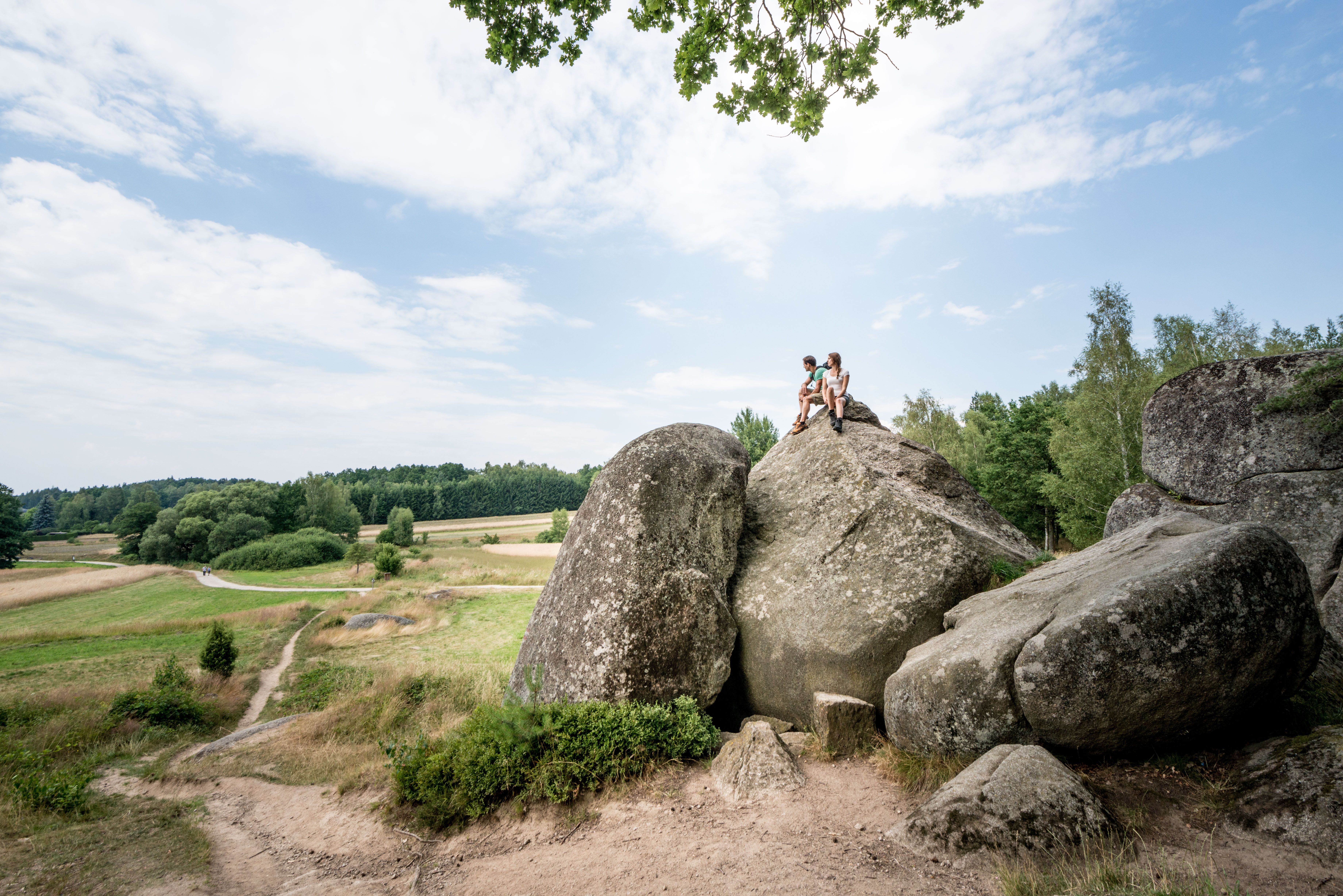 Two people are sitting on a large rock in a rural landscape with meadows and trees.