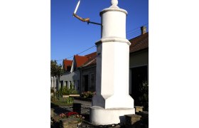 A white pillory with a wooden hand holding a sword in front of a house with red roofs.