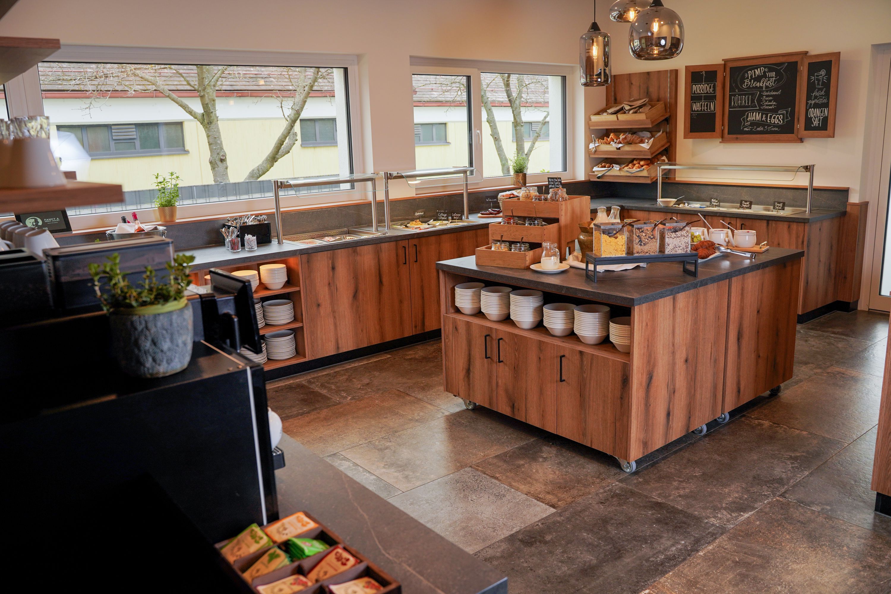 Breakfast room with a buffet, wooden counter, bowls, cereal, pastries, and large windows. The title “Egino” does not clearly correspond to the room shown.