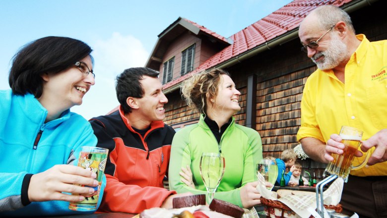 Three people sit laughing at a table in front of a hut while a man in a yellow shirt serves drinks.