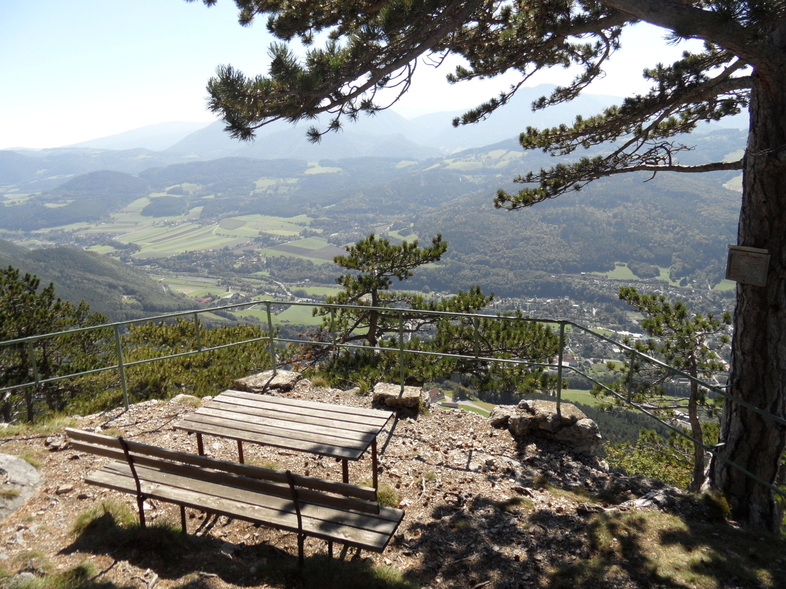 Viewpoint with bench and table, surrounded by trees, with a view of a valley and mountains in the background.