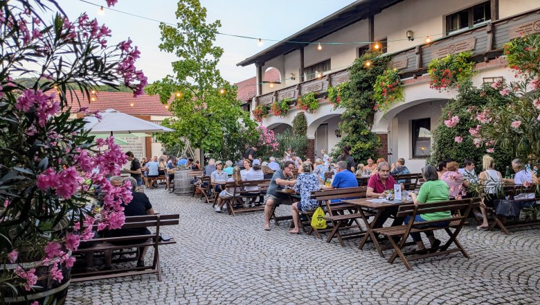 A cozy guest garden with wooden tables and benches, surrounded by flowering plants and fairy lights. People sit and enjoy the atmosphere.