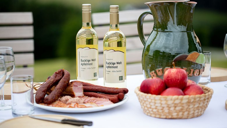 Table with cider, sausage platter, apples, wooden board, knife and glasses.