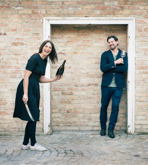 Two people in front of a brick wall, one holding a bottle and a knife, the other a glass.