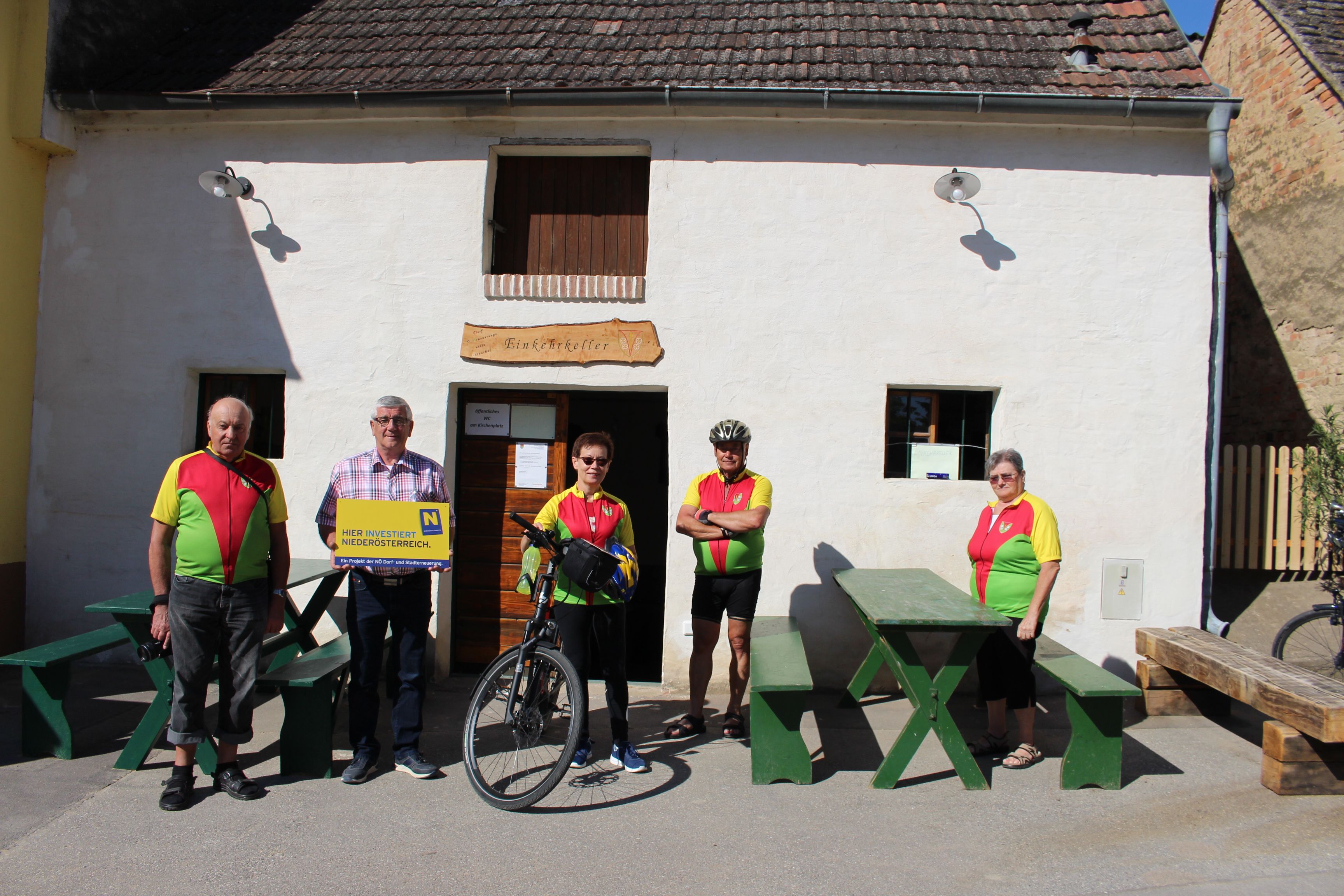 A group of five people is standing in front of the Ottenthal restaurant. Two of them are wearing cycling jerseys, and one is holding a bicycle. A sign highlights investments in Lower Austria.