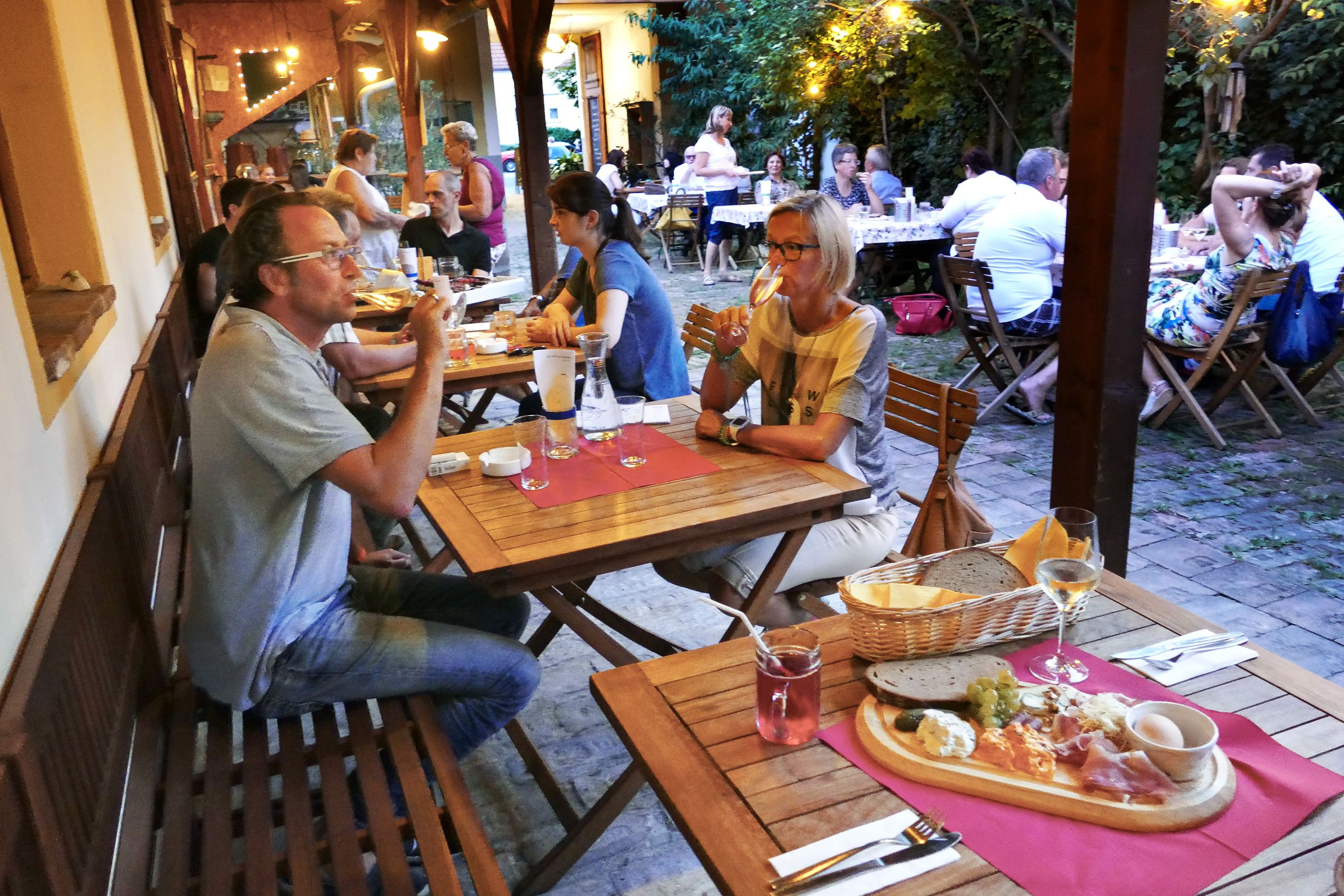People sit at wooden tables in a cozy outdoor area and enjoy food and drinks.