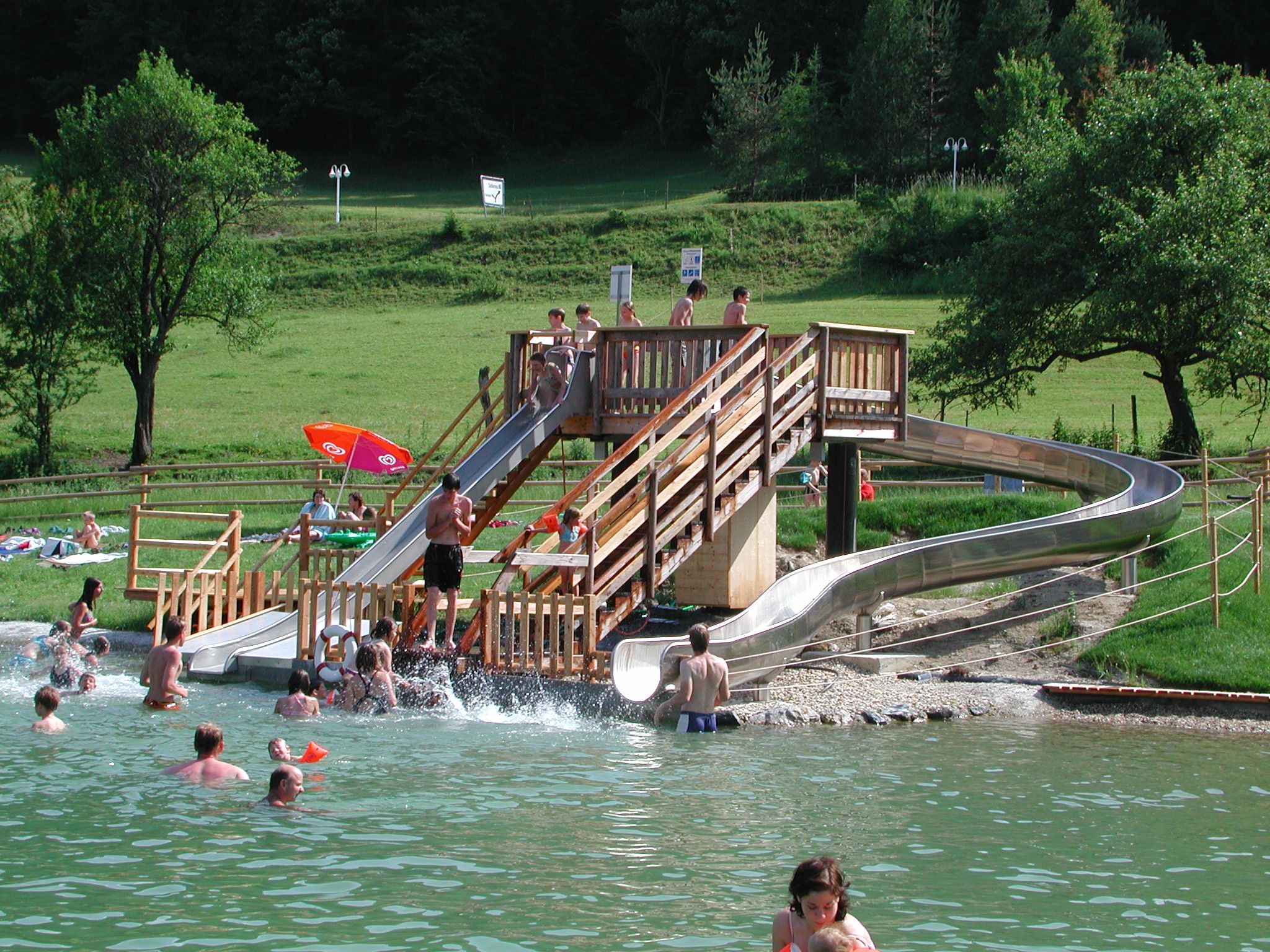 People enjoy a water slide in a natural pool with green surroundings.