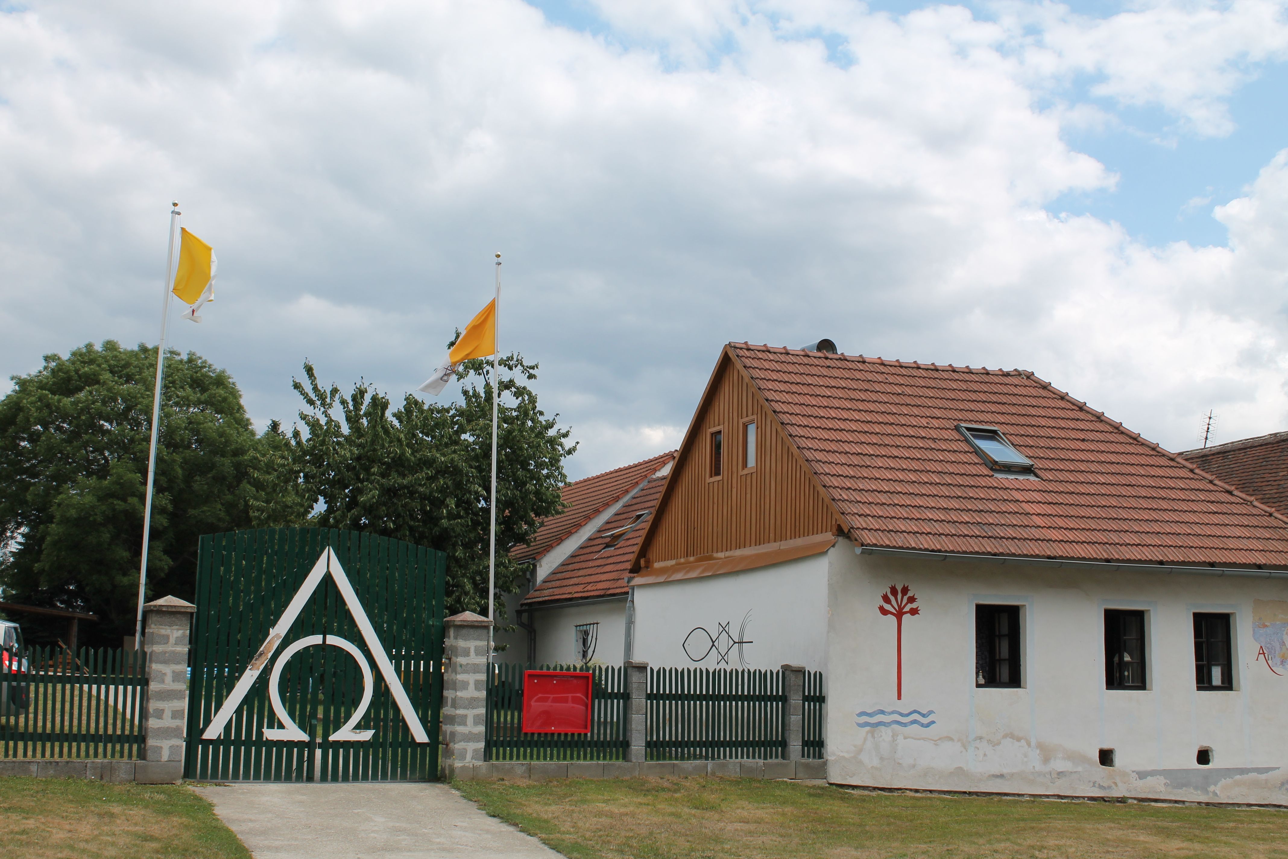 A house with red roof tiles, a green fence with the Alpha Omega symbol, two yellow flags and murals.