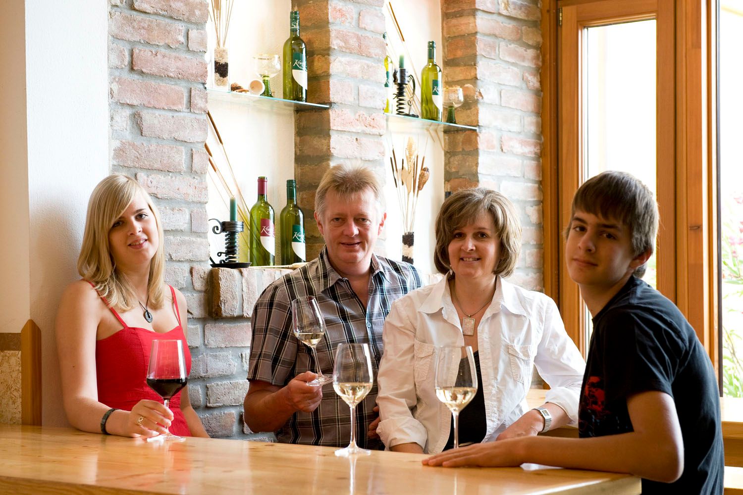 Four people sit at a table with wine glasses, wine bottles on shelves in the background.