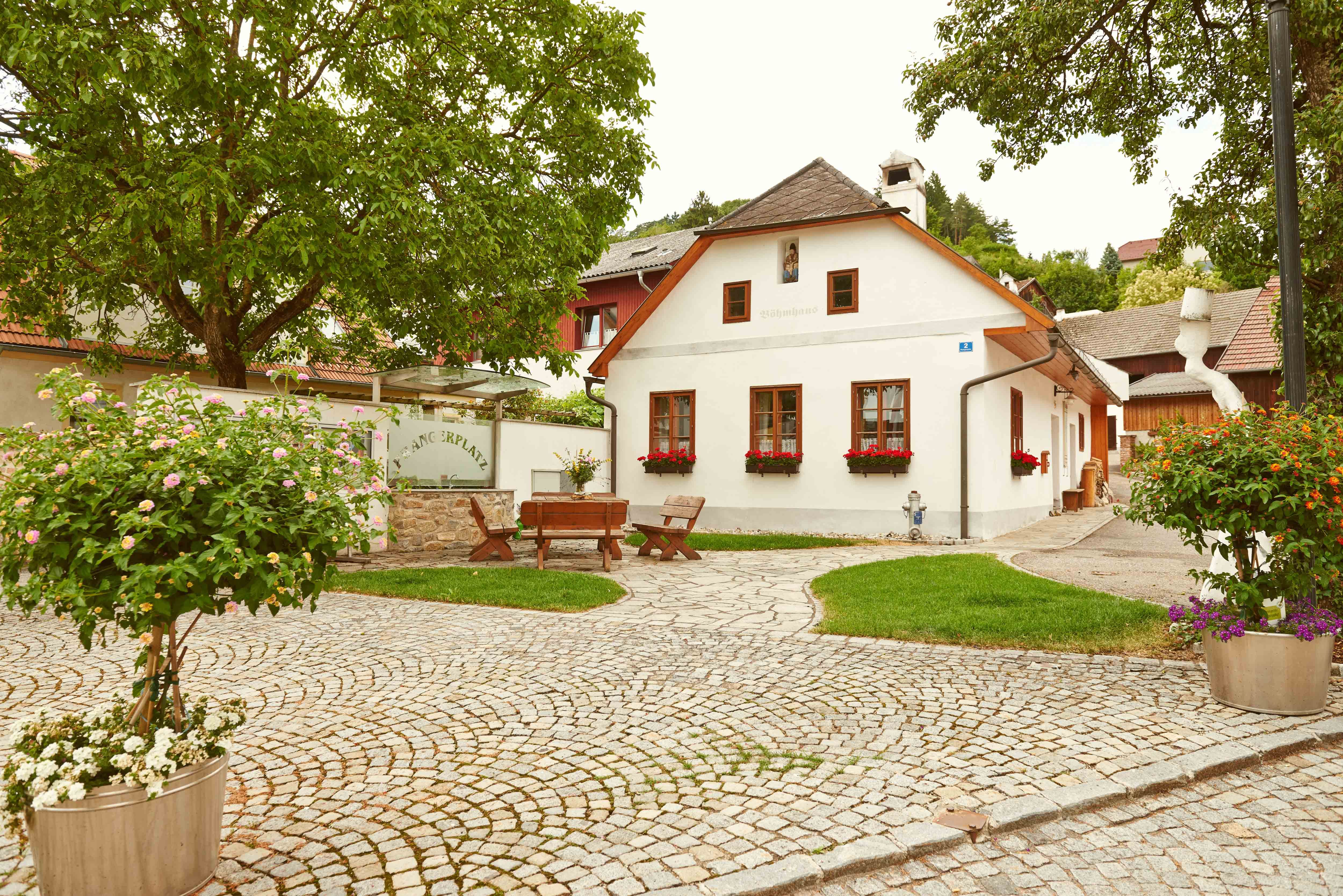 A picturesque square with cobblestones, a small white house with red shutters and flowers, surrounded by trees and plants.