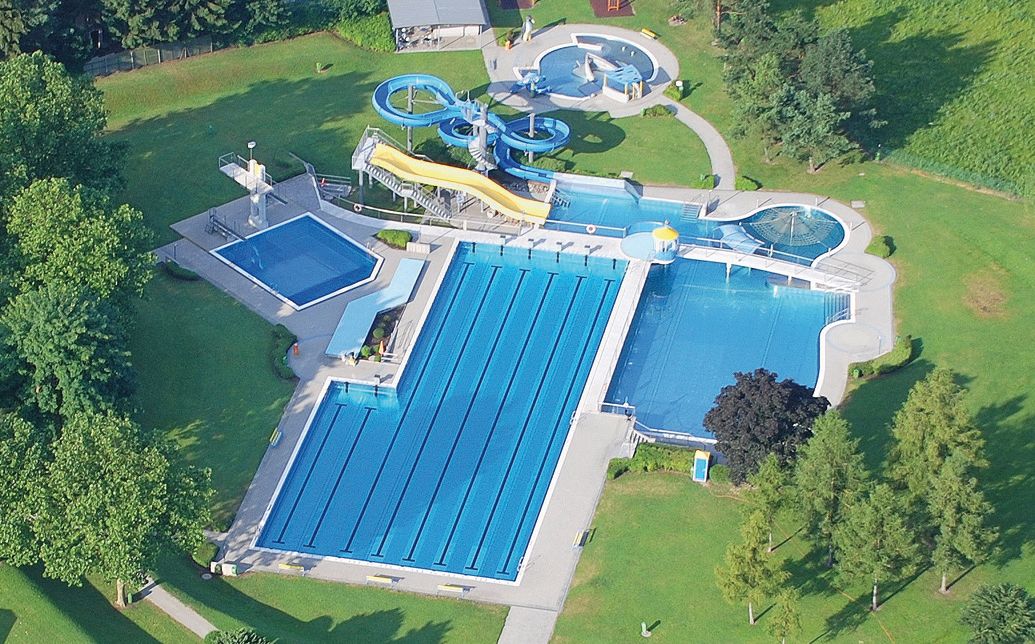 Aerial view of an outdoor pool with several pools and water slides, surrounded by green trees and lawns.