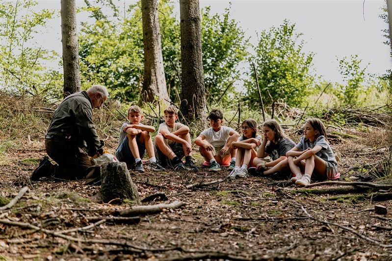 An elderly man speaks to a group of children in the forest.