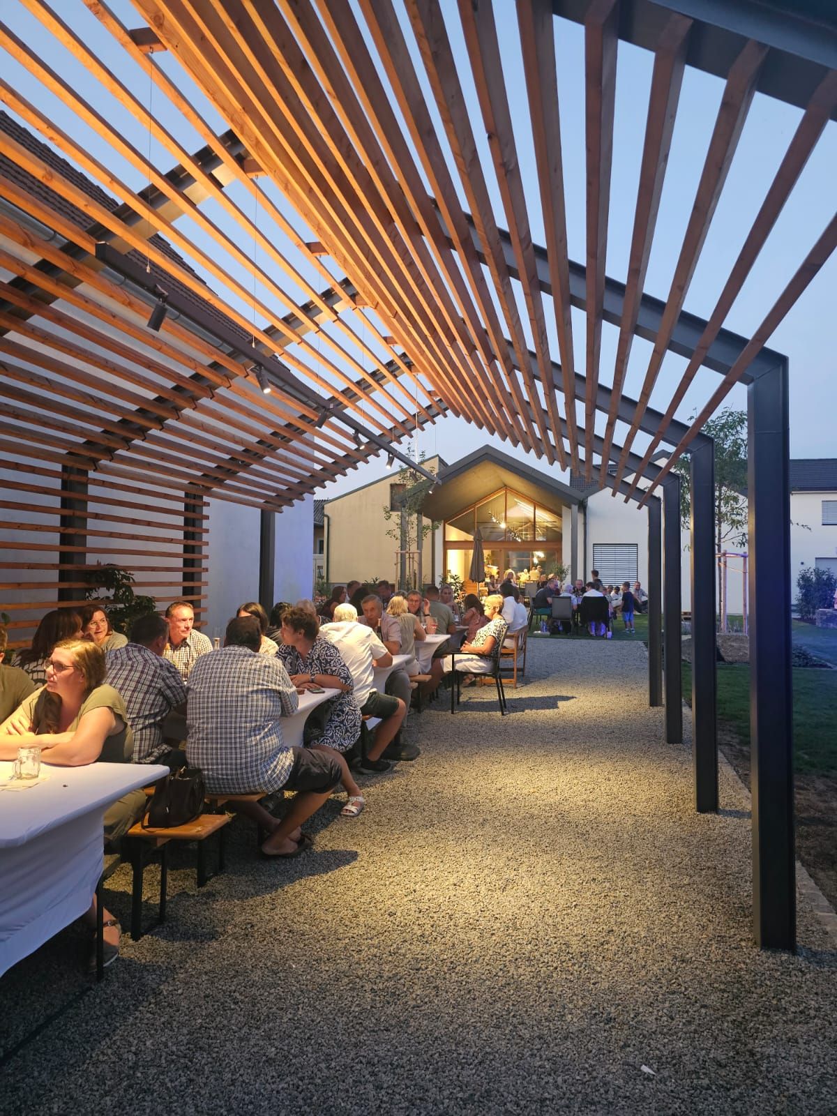 People sit at long tables under a wooden pergola at dusk.