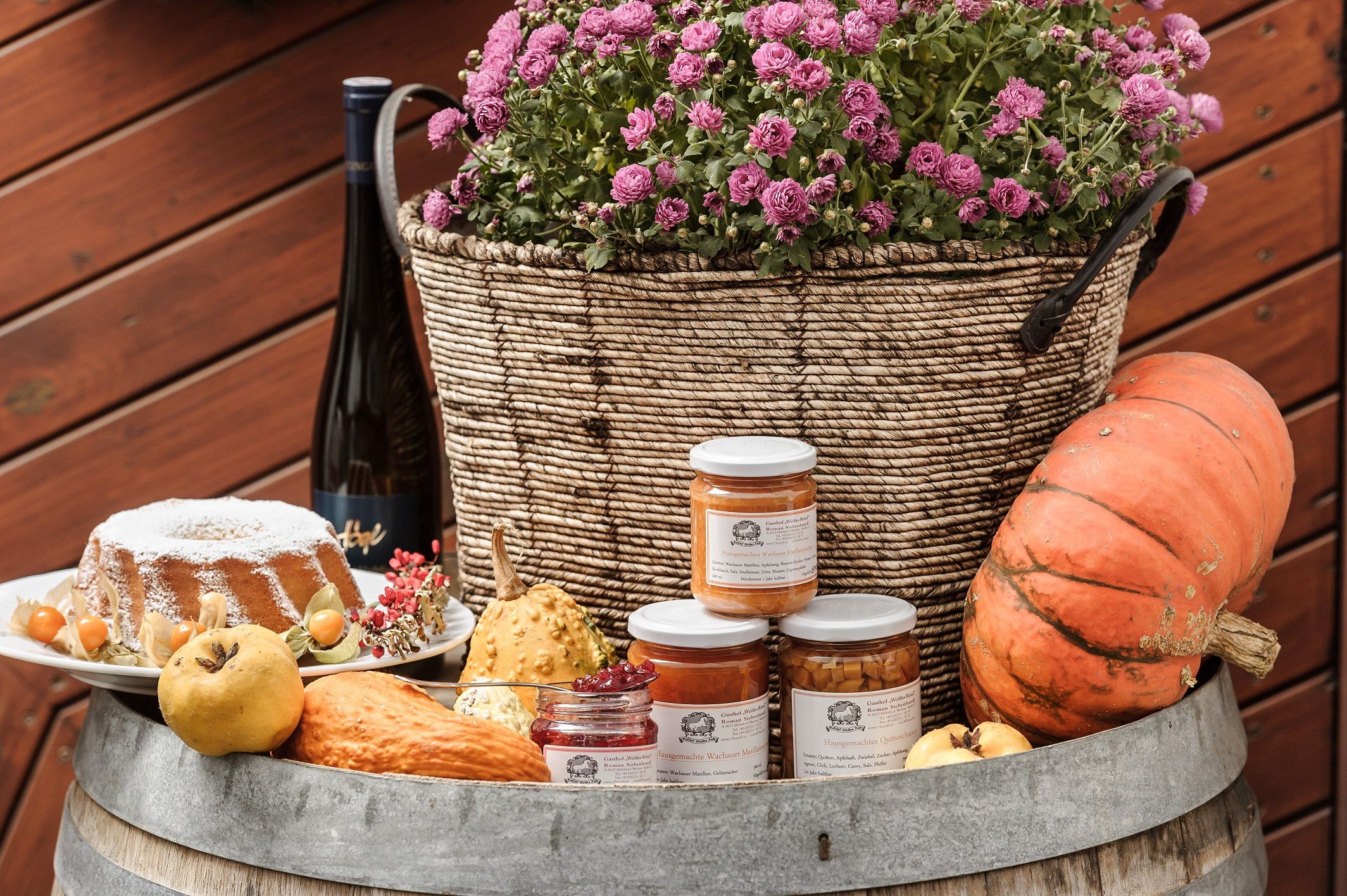 An arrangement of homemade specialties with pumpkin, jam, cake and flowers in a basket on a barrel.
