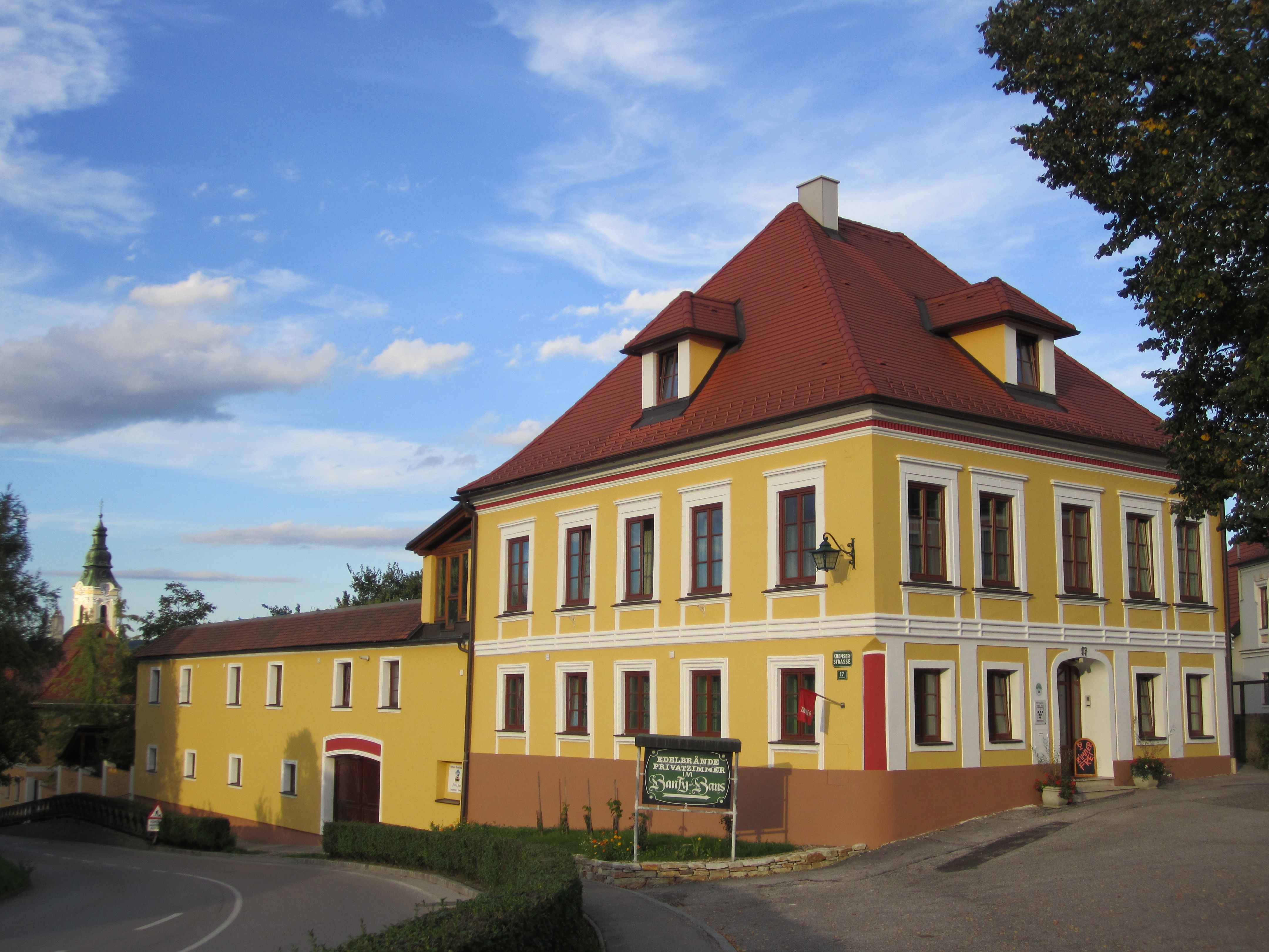 Yellow two-storey building with red roof and white window frames, next to it a smaller building. A church spire in the background.