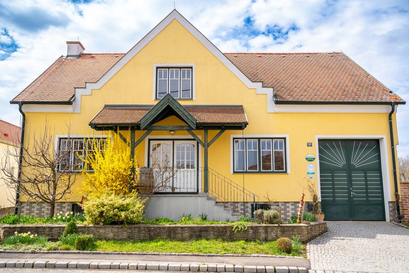 Yellow house with red roof and green gate, surrounded by garden and flowering shrubs.