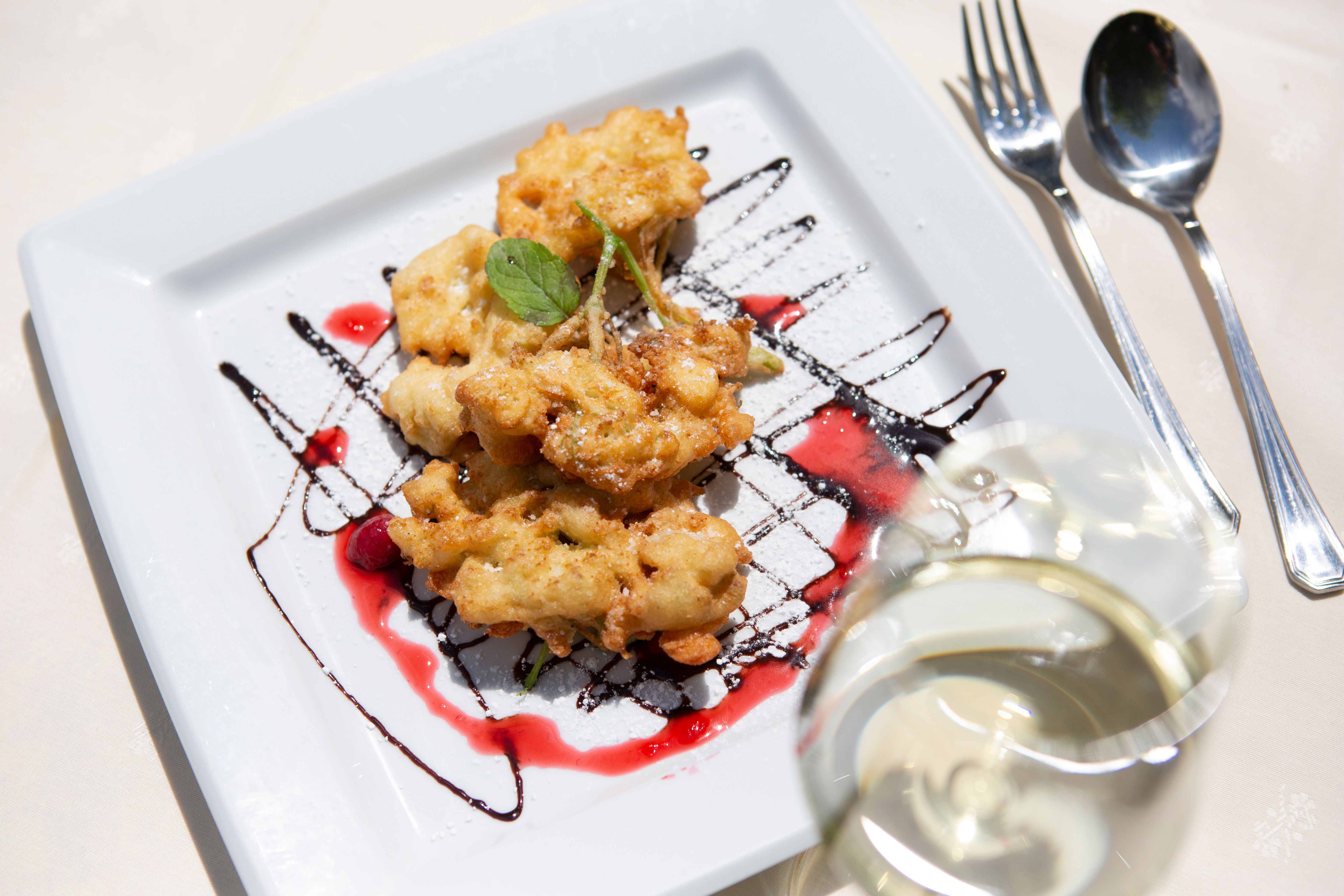 A plate of deep-fried flowers, garnished with chocolate and fruit sauce, on a white plate. A glass of white wine stands next to it.