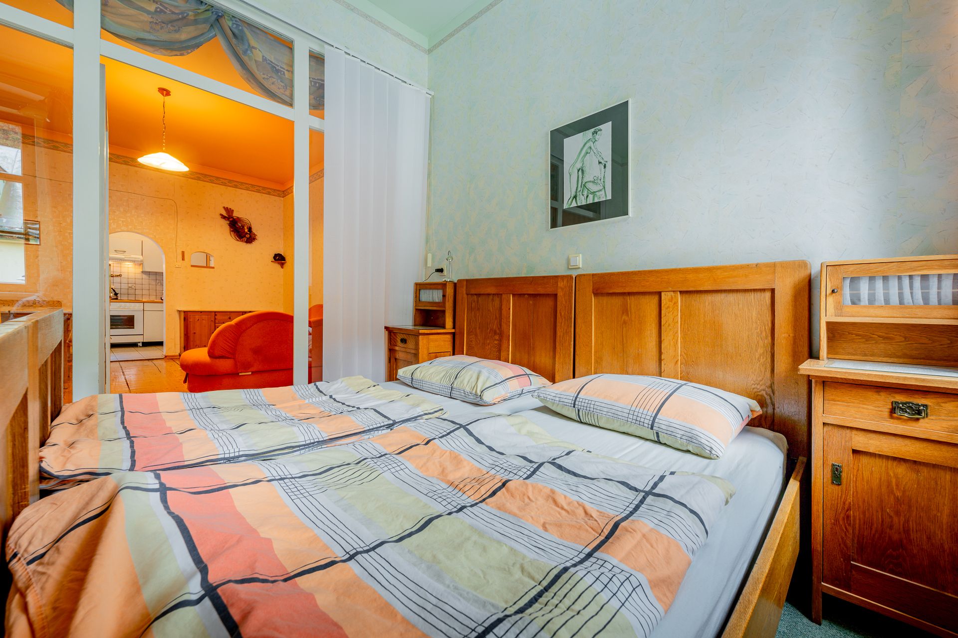 Bedroom with double bed, wooden furniture and view into a living room with orange lighting.