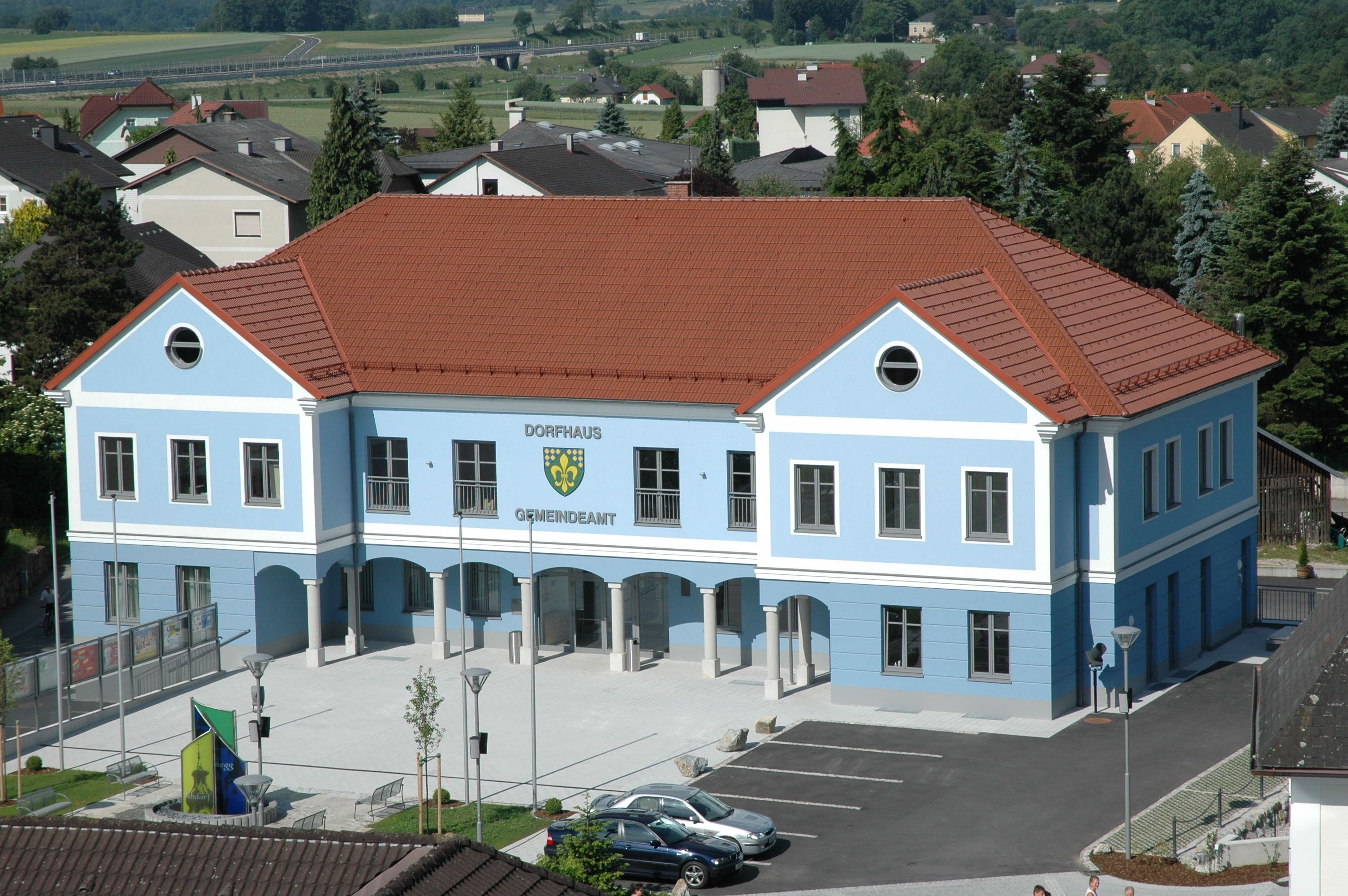 Blue building with a red roof, labeled Dorfhaus Gemeindeamt, surrounded by houses and trees.