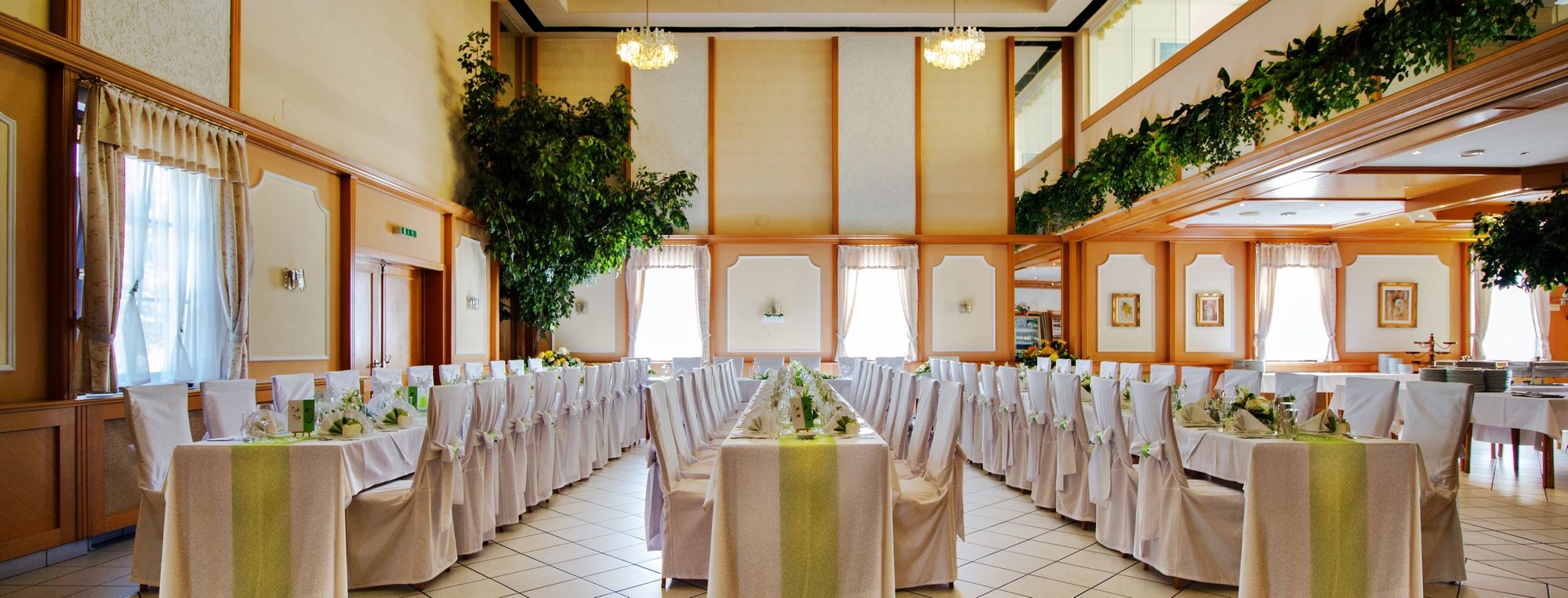 Elegant banquet hall with laid tables and white chairs, decorated with plants and chandeliers.