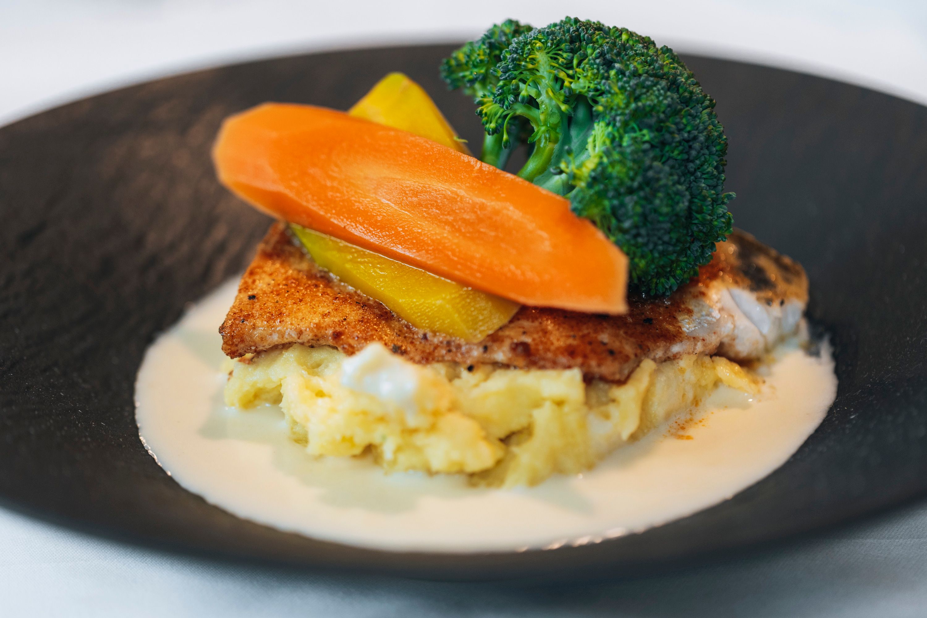 A dish of fried pike-perch on mashed potatoes, garnished with broccoli, carrots and yellow peppers, served on a black plate.
