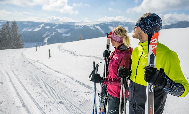 Two cross-country skiers on a snow-covered trail with a mountain panorama in the background.