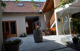 A cozy courtyard with wooden furniture, plants and a wine barrel. A building with red roof tiles and a wooden staircase in the background.