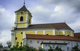 Yellow church with tower and red roofs against a cloudy sky.