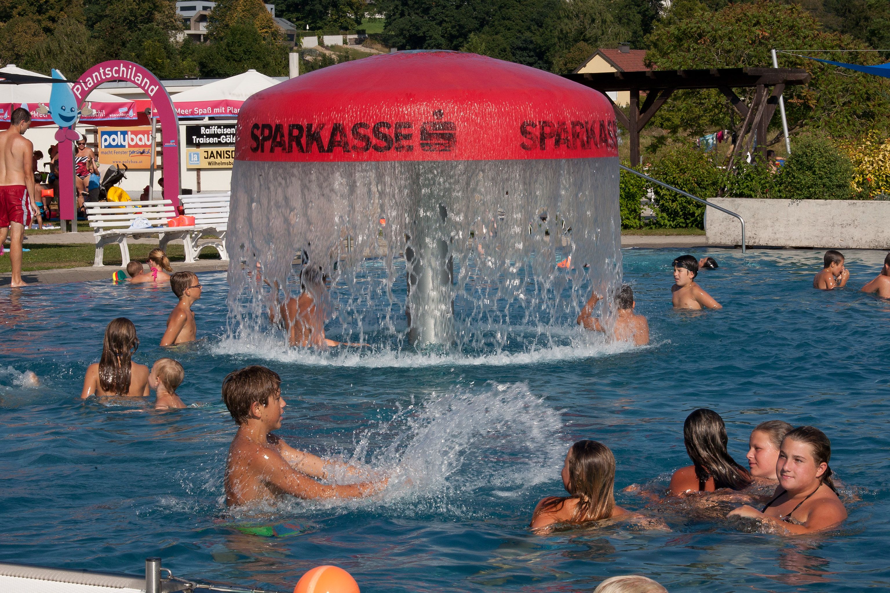 People swimming in an outdoor pool with a large water bubbler in the middle.