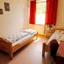 A simply furnished room with two single beds, a window with red curtains and a small table and chair.