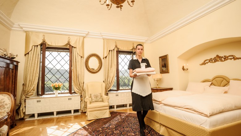 An elegant guest room with a view of the Danube, decorated in warm tones. A person in uniform holds towels.