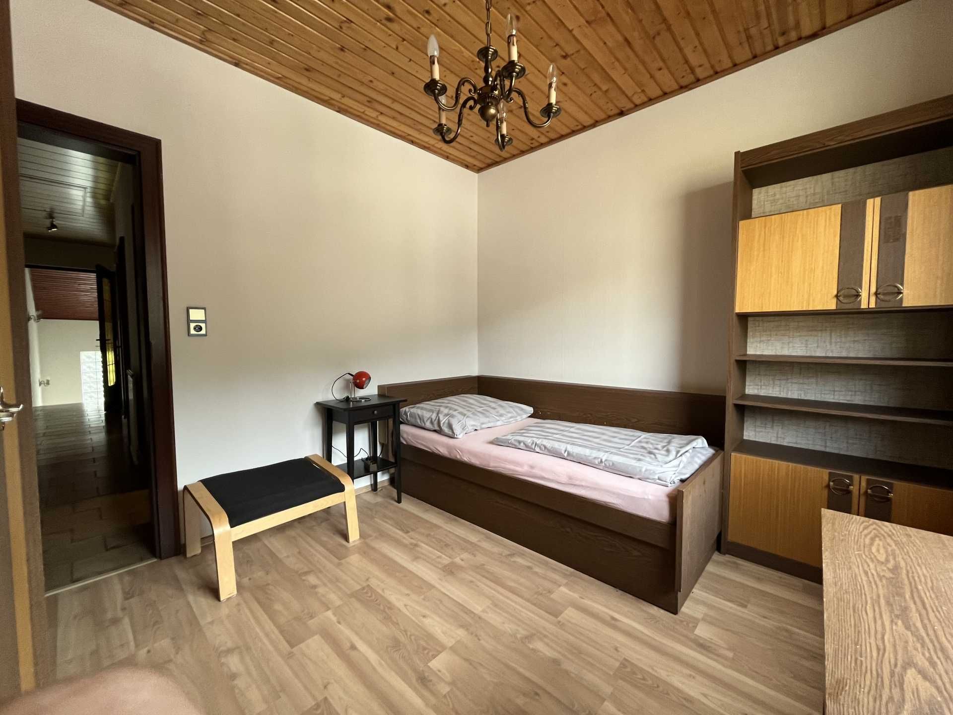 A simply furnished bedroom with single bed, wooden ceiling, shelf and small table with lamp.