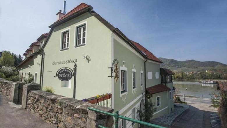 A green guest house with red roofs on the riverbank, surrounded by hills and flowering plants.