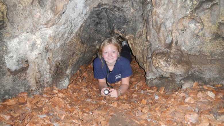 A child lies in a cave on leaves and holds a flashlight.