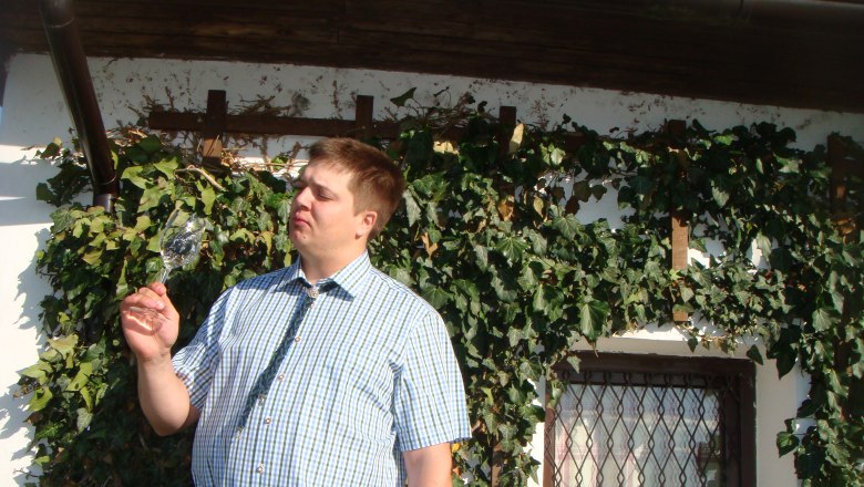 Man in plaid shirt looks at a wine glass in front of a wall covered with ivy.